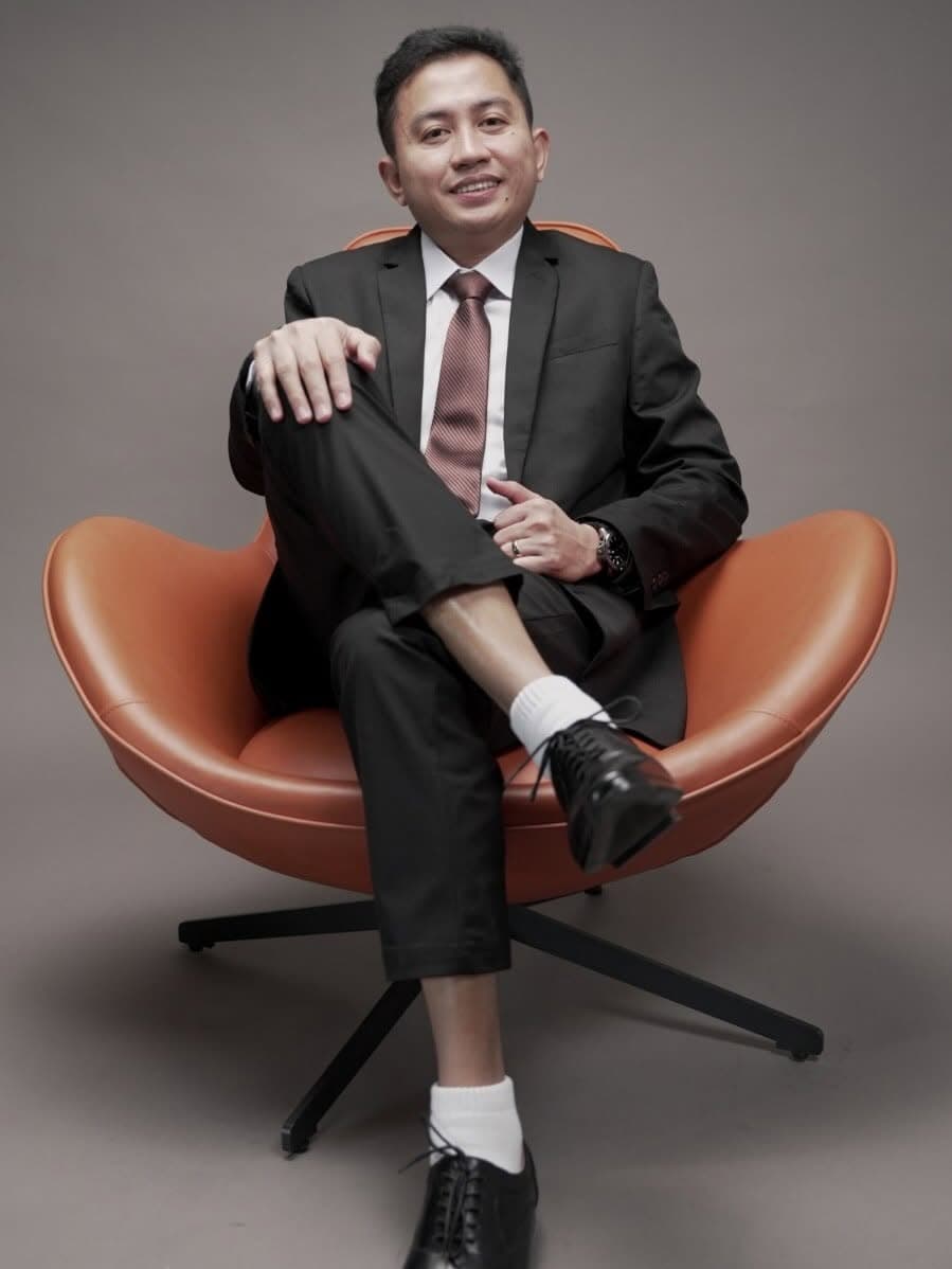 Mark Anthony Ragus, Founder and CEO of NextStack Training & Consulting Inc., seated in an orange lounge chair wearing a dark suit and tie.