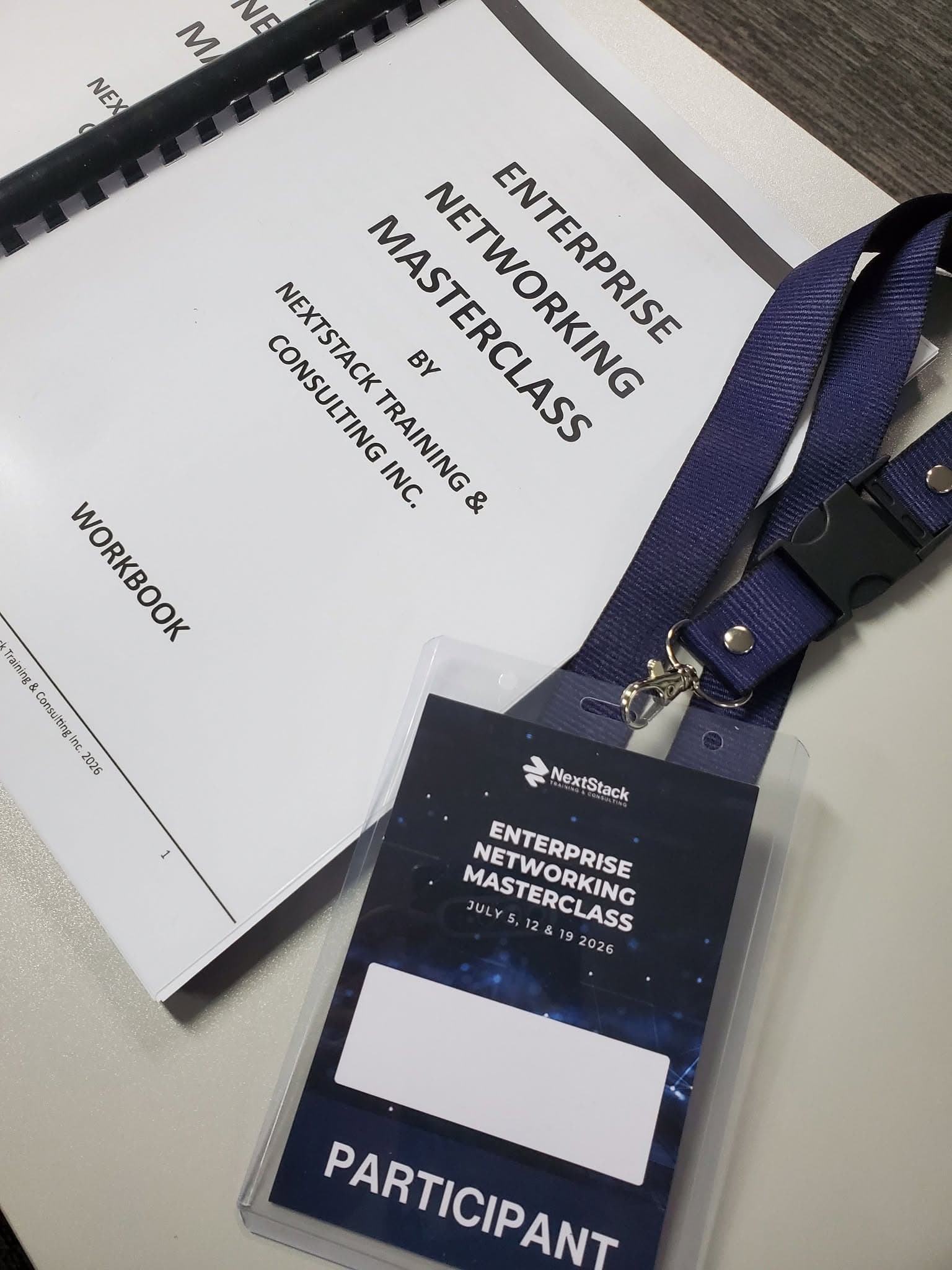 A spiral-bound Enterprise Networking Masterclass workbook by NextStack Training & Consulting Inc. lying beside a participant lanyard and badge.