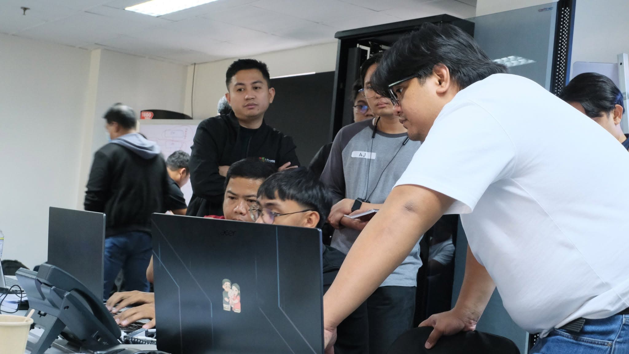 An instructor leans over a participant's laptop to guide a configuration while several other participants watch over their shoulders. A server rack and IP desk phones are visible in the room.