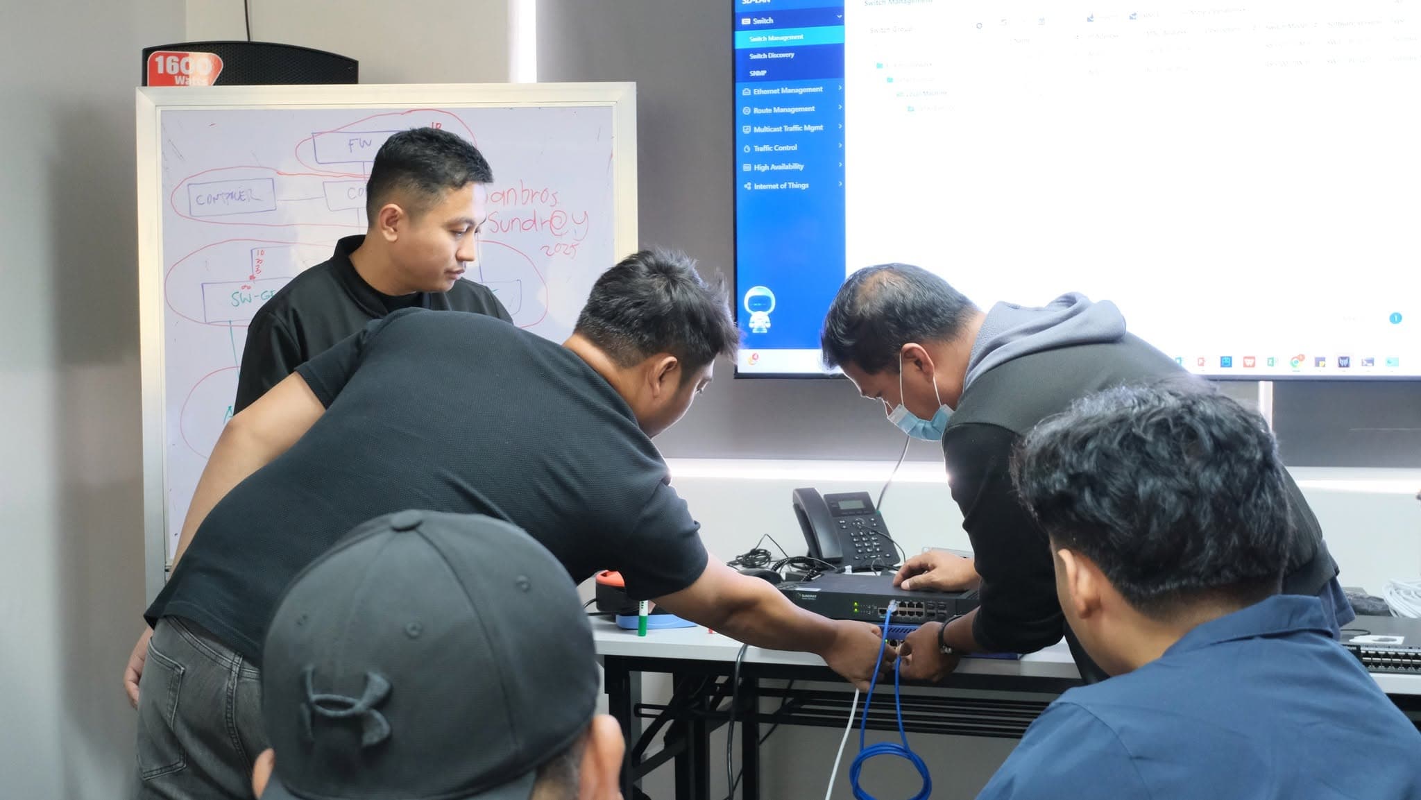 Participants gathered around a bench, one patching a blue network cable into a managed switch. A switch management interface is on the screen behind them and a hand-drawn network topology covers the whiteboard.