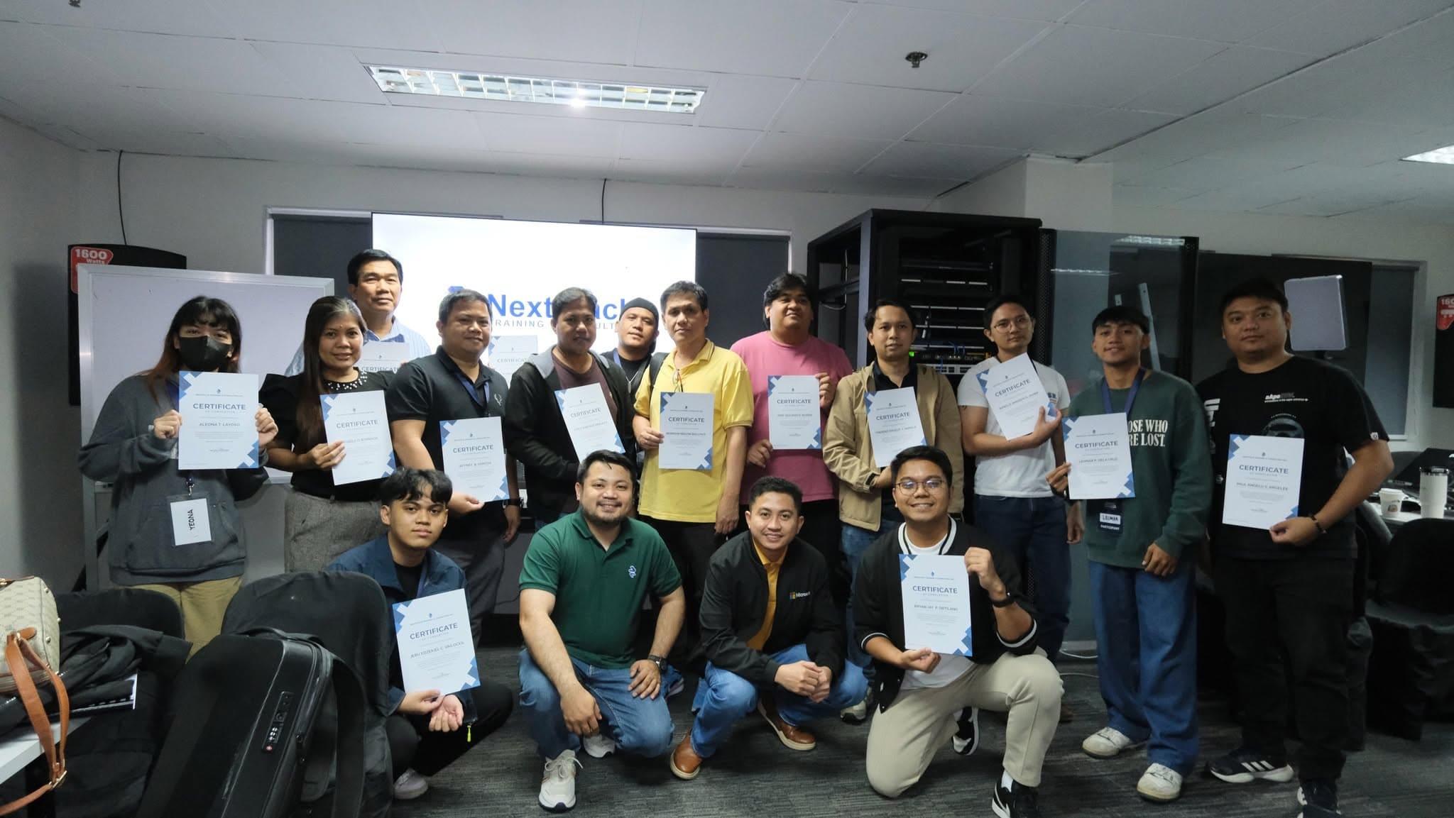 Participants posed together holding certificates of completion, with the NextStack logo projected on the screen behind them and a server rack to the side.