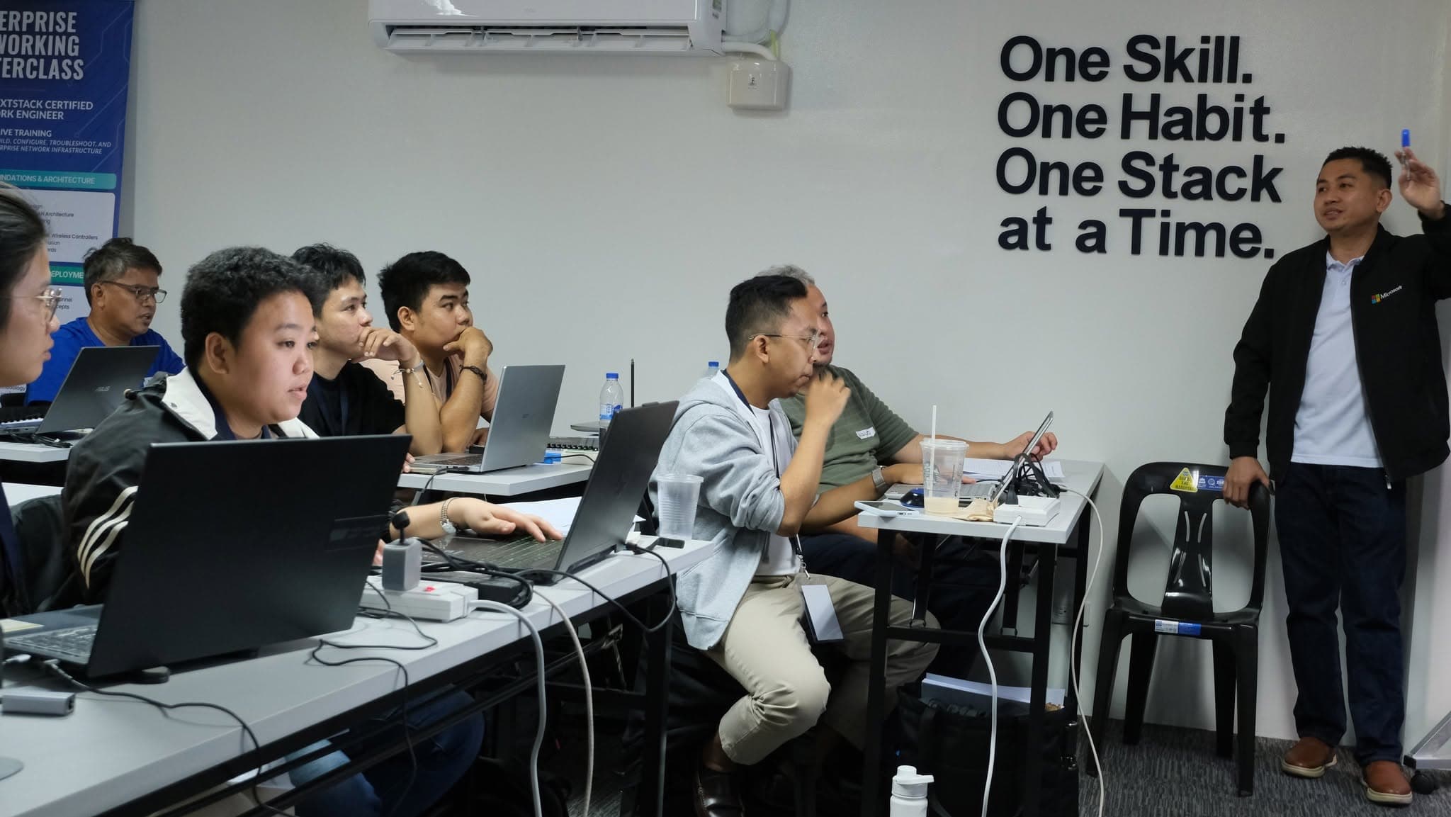 An instructor stands at the front of the NextStack training room mid-explanation while participants follow along at laptops. Large lettering on the wall behind reads "One Skill. One Habit. One Stack at a Time."
