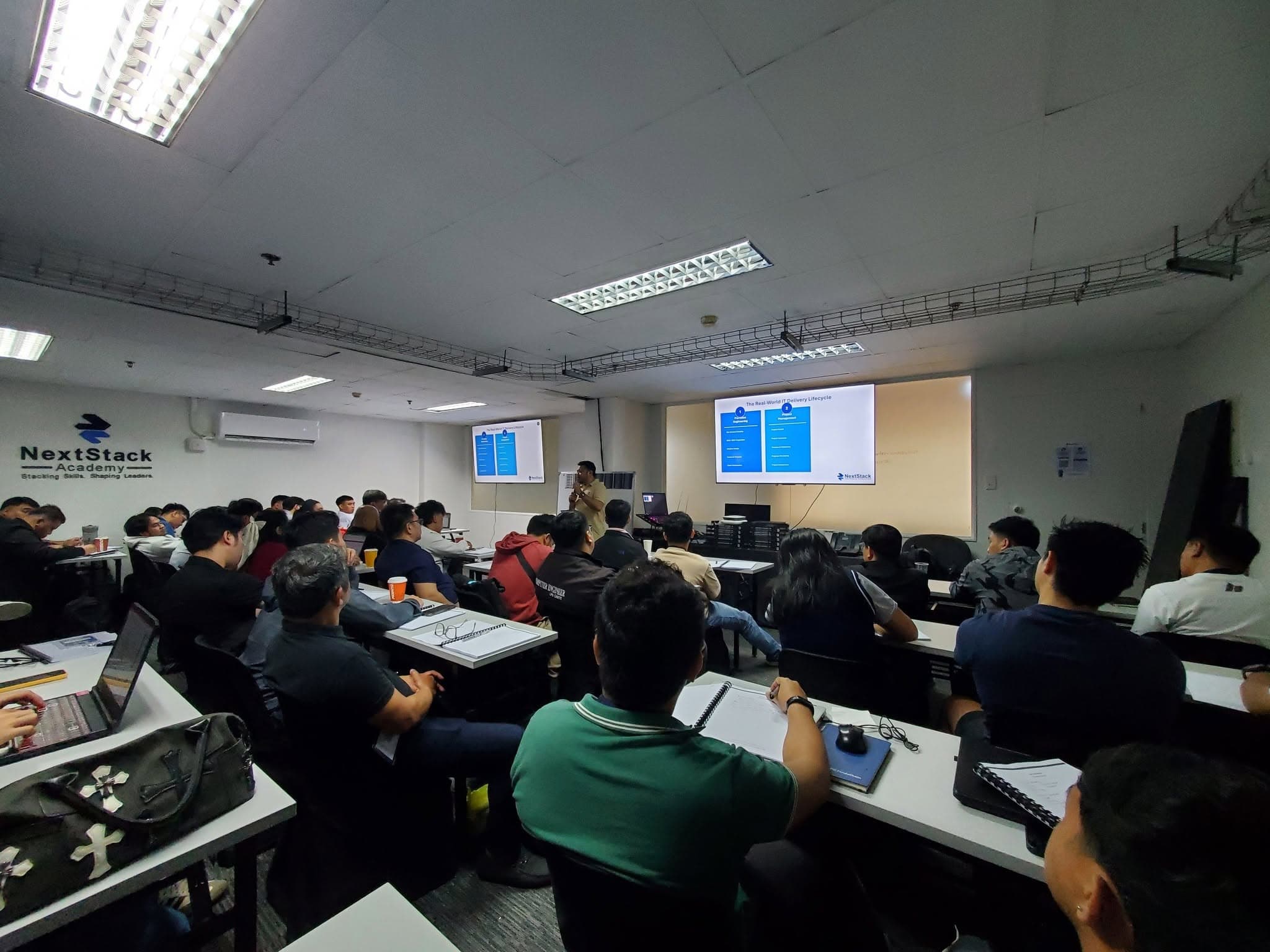 A full classroom of participants facing a presentation slide titled "The Real-World IT Delivery Lifecycle", covering presales engineering and project management. The NextStack Academy wall sign is on the left.