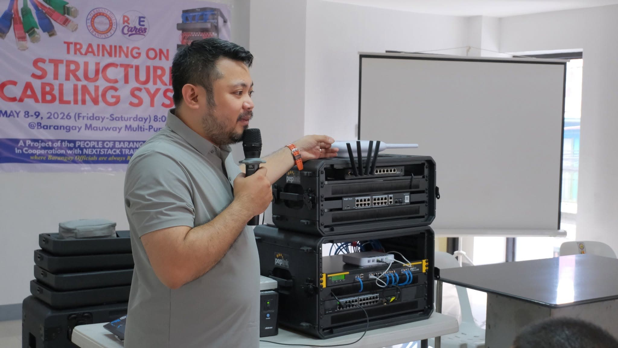 An instructor speaks into a microphone beside two stacked portable equipment racks holding switches, a router and a wireless access point. A tarpaulin behind him reads "Training on Structured Cabling System".