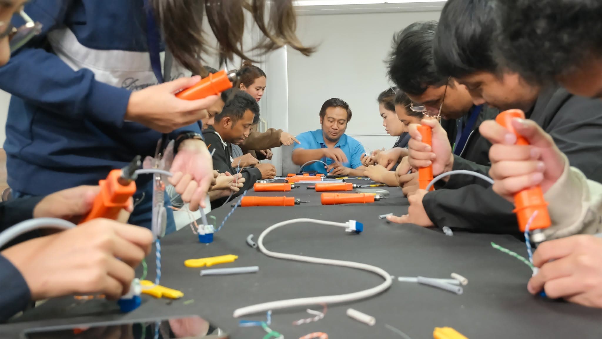 Participants seated along both sides of a long table each work an orange impact punch-down tool onto keystone jacks, while the instructor at the head of the table guides them.